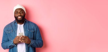 Yakışıklı Afro-Amerikalı adam akıllı telefon kullanıyor, kameraya gülümsüyor, pembe arka planda duruyor..