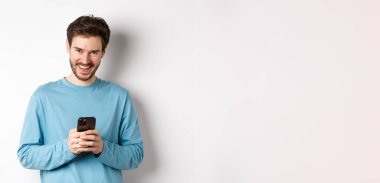 Akıllı telefon kullanan, gülen, kameraya gülümseyen, beyaz arka planda duran yakışıklı bir adam..