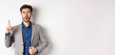 Heyecanlı takım elbiseli işadamı iyi fikir sunuyor, Eureka işaretinde parmağını kaldırıyor ve komik görünüyor, öneriler söylüyor, beyaz arka planda duruyor..