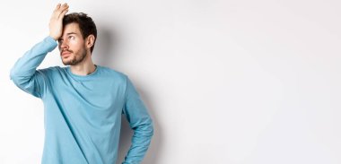 Annoyed young man with beard, roll eyes and making facepalm, pissed-off with something, standing irritated on white background.