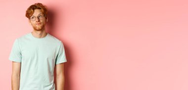 Sakallı, tişörtlü ve gözlüklü, canı sıkkın, pembe arka planda duran genç bir kızıl adam..