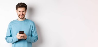 Gülümseyen modern adam akıllı telefon kullanıyor ve kameraya mutlu bakıyor. Beyaz arka planda cep telefonu olan kazaklı bir adam..