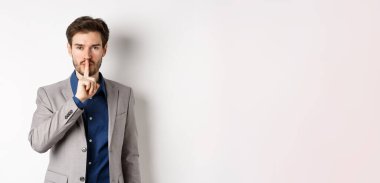 Yakışıklı maço takım elbiseli adam kameraya şşş yapıyor, parmak dudağa basıyor, sesini alçalt, sırrını söyle, beyaz arka plana karşı duruyor..