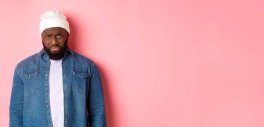 Hüsrana uğramış ve üzgün siyah adam, somurtuyor ve mızmızlanıyor, bozuk suratla kameraya bakıyor, pembe arka planda duruyor..