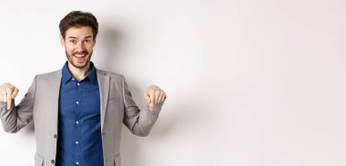 Hey, şuna bak. Takım elbiseli mutlu işadamı parmaklarını aşağı uzatıp gülümsüyor, etkinliğe davet ediyor, bağlantıyı gösteriyor, beyaz arka planda duruyor..