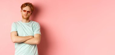 Turizm ve tatil konsepti. Güneş gözlüklü, göğsünde çapraz kolları olan, pembe arka planda duran ve kameraya ciddi ciddi bakan arsız kızıl saçlı adam..