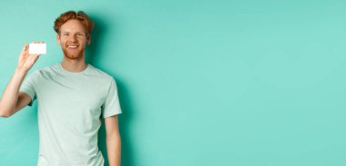 Alışveriş konsepti. Plastik kredi kartı gösteren, gülümseyen, turkuaz arka planda duran, tişörtlü yakışıklı kızıl adam..