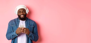 Yakışıklı siyahi adamın kulaklıklı, müzik dinleyen ve cep telefonu kullanan, kameraya gülümseyen, pembe arka planlı bir resmi..