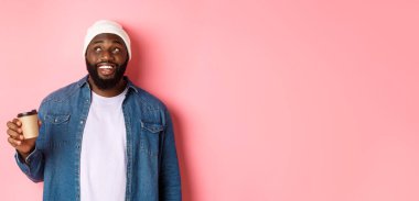 Yakışıklı, modern siyahi adam paket kahve içiyor, gülümsüyor ve kameraya tatmin olmuş görünüyor, pembe arka planda duruyor..