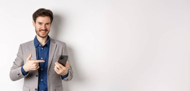 Takım elbiseli başarılı adam cep telefonunu işaret ederek, mobil uygulamayı tanıtarak, beyaz arka planda durarak.
