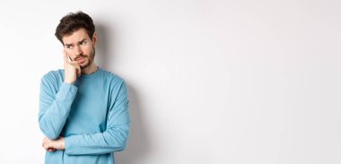 Tişörtlü sakallı endişeli genç adam, dalgın dalgın bakıyor ve düşünüyor, beyaz arka plana karşı sorunlu duruyor. Boşluğu kopyala