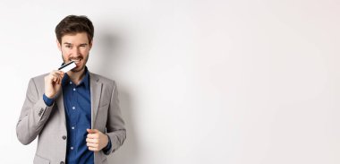 Başarılı takım elbiseli genç işadamı kredi kartını ısırıyor ve beyaz arka planda arsız görünüyor..
