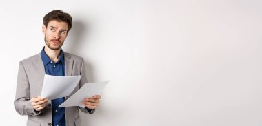 Takım elbiseli bir işadamı gazetelere bakıyor, işte belgeler okuyor ve düşünüyor, logoya bakıyor, beyaz arka planda meşgul duruyor..