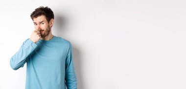 Tişörtlü iğrenç genç adam burnunu kapadı, kötü kokulu bir şeye baktı, beyaz arka planda duruyordu..