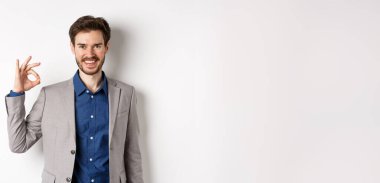 Gülümseyen başarılı takım elbiseli işadamı tamam işareti gösteriyor, tatmin olmuş görünüyor, beyaz arka plana karşı duruyor..