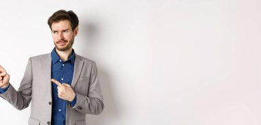 Takım elbiseli hoşnutsuz işadamı şüpheci bir sırıtışla sol tarafa bakıyor, pankarttan hoşlanmıyor, onaylanmayan promosyon teklifi, beyaz arka planda duruyor..