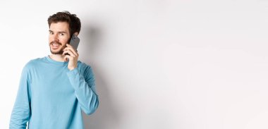 Teknoloji konsepti. Genç erkek model cep telefonuyla konuşuyor, akıllı telefondan birini arıyor ve gülümsüyor, beyaz arka planda duruyor..