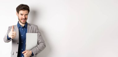 Başarılı bir iş adamı şık bir takım elbiseyle dizüstü bilgisayar taşır ve kameraya kendinden emin bakar, onaylı, beyaz arka planda başparmak kaldırır..