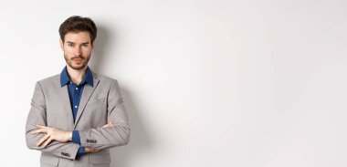 Profesyonel gibi görünen kendine güvenen genç adam, göğsünde çapraz kollar, beyaz arka planda duruyor..