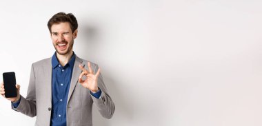 E-ticaret ve online alışveriş konsepti. Başarılı iş adamı akıllı telefondan para kazanıyor, boş telefon ekranı ve tamam işareti gösteriyor, kameraya mutlu bir şekilde göz kırpıyor..