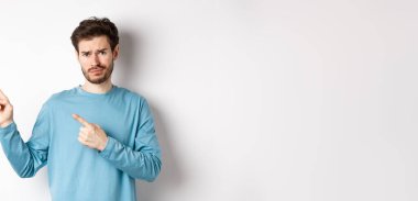 Şüpheci genç adam sakallı, tatminsiz, somurtkan, sol üst köşedeki logoyu işaret eden şüpheli, beyaz arka planda üzgün duruyor..