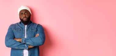 Hipster bereli, hayal kırıklığına uğramış Afro-Amerikan bir adam, kameraya hoşnutsuzca bakıyor, göğsünde çapraz kollar, pembe arka planda duruyor..