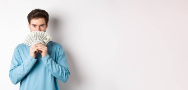 Finans konsepti. Gülümseyen genç adam paranın arkasında yüzünü örtüyor, dolarları gösteriyor ve mutlu görünüyor, beyaz arka planda duruyor..