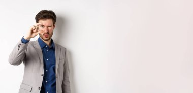 İş adamı küçük bir şey görmeye çalışıyor, parmaklarıyla küçük bir şey gösteriyor, beyaz arka planda takım elbise içinde duruyor..