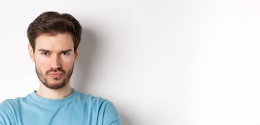 Suratsız, somurtkan ve gücenmiş suratlar yapan, beyaz arka planda üzgün duran huysuz genç adamın yakın çekimi..