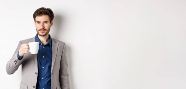 Takım elbiseli yakışıklı işadamı kahve ya da ofis kupasından çay içiyor, beyaz arka planda duruyor..