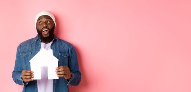 Emlak konsepti. Yakışıklı Afro-Amerikalı adam evini düşlüyor, ev modelini tutuyor ve sol üst köşeye bakıyor, resim dairesi, pembe arka plan..