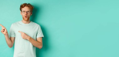 Gözlüklü, tişörtlü, sol elini gösteren ve tanıtım teklifini gösteren, turkuaz arka planda duran şaşkın kızıl saçlı adam..