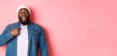 Hipster bereli şaşkın siyah adam kendini işaret ediyor, kameraya şaşkın şaşkın bakıyor, pembe arka planda duruyor..