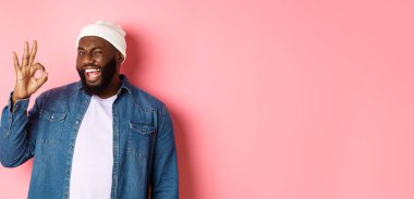 Mutlu gülümseyen Afrikalı Amerikalı adam pembe arka planda duran iyi bir işaret, onay ve övgü veriyor..