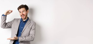 Takım elbiseli yakışıklı bir satıcı, büyük gelir elde ediyor, kameraya hevesle gülümsüyor, beyaz arka planda duruyor..