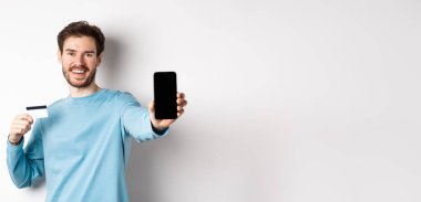 E-ticaret ve alışveriş konsepti. Gülümseyen beyaz adam plastik kredi kartı ve boş akıllı telefon ekranı gösteriyor. Çevrimiçi uygulama öneriyor..