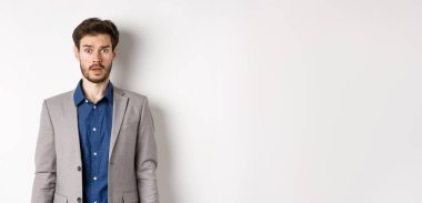 Şok olmuş takım elbiseli girişimci, endişeli ve şaşkın bir şekilde kameraya bakıyor, beyaz arka planda duruyor..