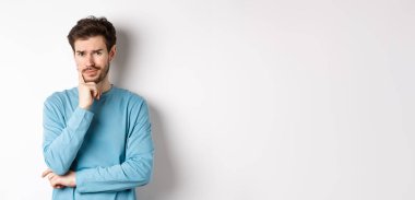 Şüpheci genç adam somurtuyor, endişeli düşünceli bakıyor, beyaz arka plan üzerinde üzgün duruyor.