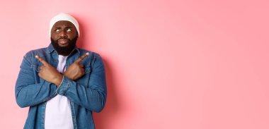 Kafası karışmış sakallı siyah bir adam, seçim yapıyor, parmaklarını yana doğru gösteriyor ve şaşkın şaşkın bakıyor, pembe arka planda duruyor..