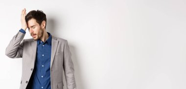 Annoyed businessman in suit facepalm with eyes closed, bothered by someone stupid, pissed-off at work, standing on white background.