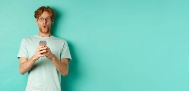 Kızıl saçlı, gözlüklü, elinde cep telefonu olan etkileyici bir genç adam kameraya hayret ediyor ve bakıyor..