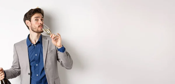 stock image Celebration and holidays concept. Handsome bearded man in suit and birthday hat holding bottle, drinking glass of champagne, standing on white background.