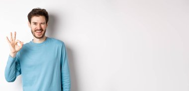 Yakışıklı, kendine güvenen, tamam işareti gösteren ve sana göz kırpan, her şeyin yolunda olduğuna emin olan, beyaz arka planda duran,.