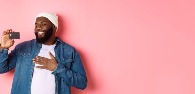 Alışveriş konsepti. Mutlu Afro-Amerikalı bir adamın resmi kredi kartından memnun görünüyor, bankayı tavsiye ediyor, pembe arka planda duruyor..