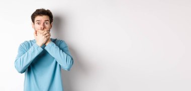 Şok olmuş genç bir adamın elleri ile ağzını kapayarak bakması, dedikoduları duyması, beyaz arka planda durması..