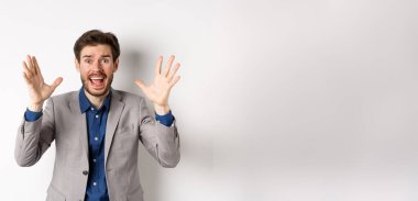 İş adamı çığlık atıyor ve panik içinde el sıkışıyor, tedirgin ve endişeli görünüyor, kameraya bağırıyor, beyaz arka planda takım elbise içinde duruyor..