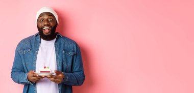 Neşeli Afro-Amerikalı adam doğum gününü kutluyor, doğum günü pastasında mumla dilek diliyor, mutlu bir şekilde gülümsüyor, pembe arka planda duruyor..