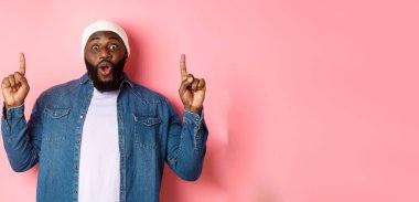 Heyecanlı ve etkilenmiş siyah adam harika bir tanıtım sergiliyor, parmaklarını logoya doğrultuyor, pembe arka planda duruyor..