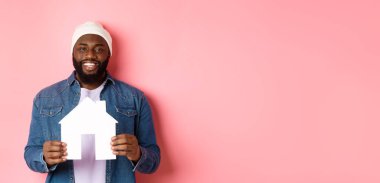 Emlak konsepti. Mutlu gülümseyen Afro-Amerikalı adam ev modelini tutuyor, daire alıyor, pembe arka planda duruyor..