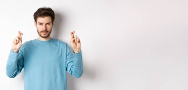 Umut dolu ve kendinden emin genç adam şans dilemek ya da dua etmek için parmaklarını çapraz yapıyor, sonuçları tahmin ediyor, beyaz arka planda duruyor..